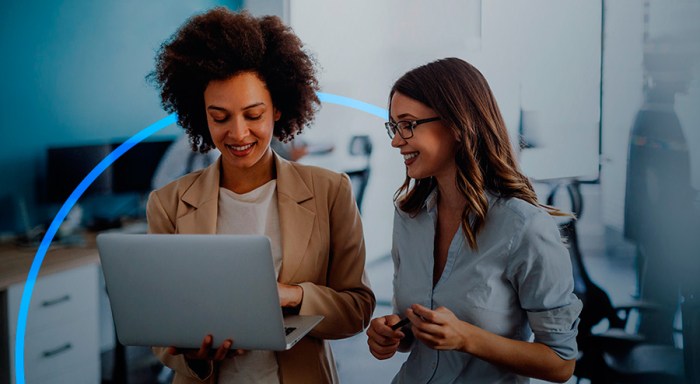 Two women standing in an office talking while they look at the laptop computer held by the woman on the left